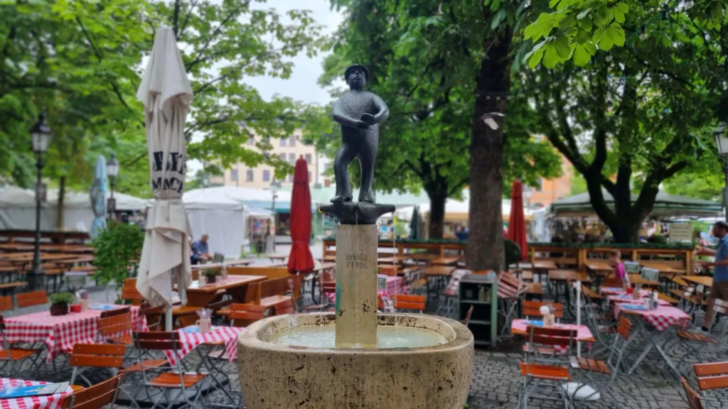 Fotografie des Weiß-Ferdl-Brunnens auf dem Viktualienmarkt umgeben von den Tischen und Stühlen des Biergartens